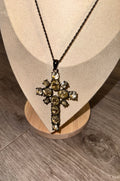 A cross-shaped Gothic Style pendant necklace with clear faceted gemstones set in a dark metal, displayed on a beige fabric jewelry bust over a wooden stand on a gray wood-look floor.