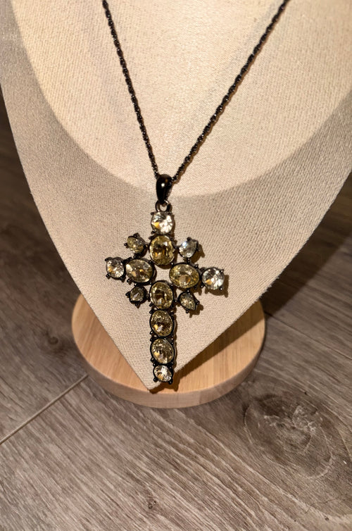 A cross-shaped Gothic Style pendant necklace with clear faceted gemstones set in a dark metal, displayed on a beige fabric jewelry bust over a wooden stand on a gray wood-look floor.