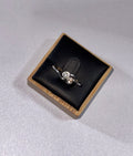 An 18 ct Gold ring with 2 diamonds  small side accents, crossovers, displayed on a black cushioned square box with a light wood edge, set against a light gray textured background.