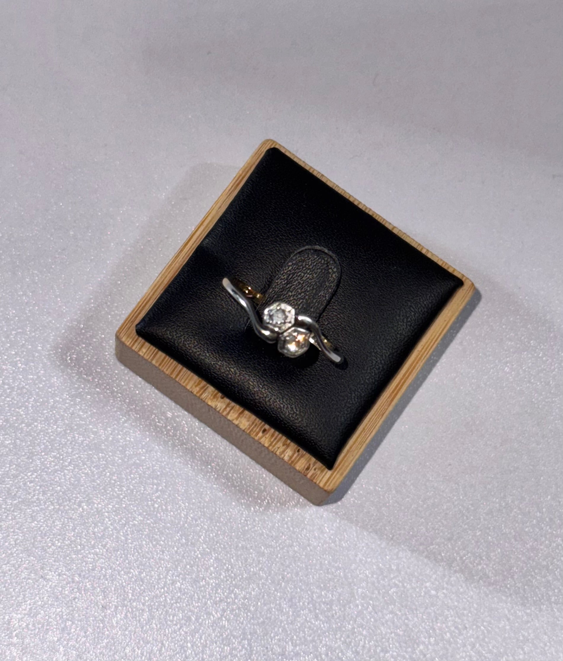 An 18 ct Gold ring with 2 diamonds  small side accents, crossovers, displayed on a black cushioned square box with a light wood edge, set against a light gray textured background.