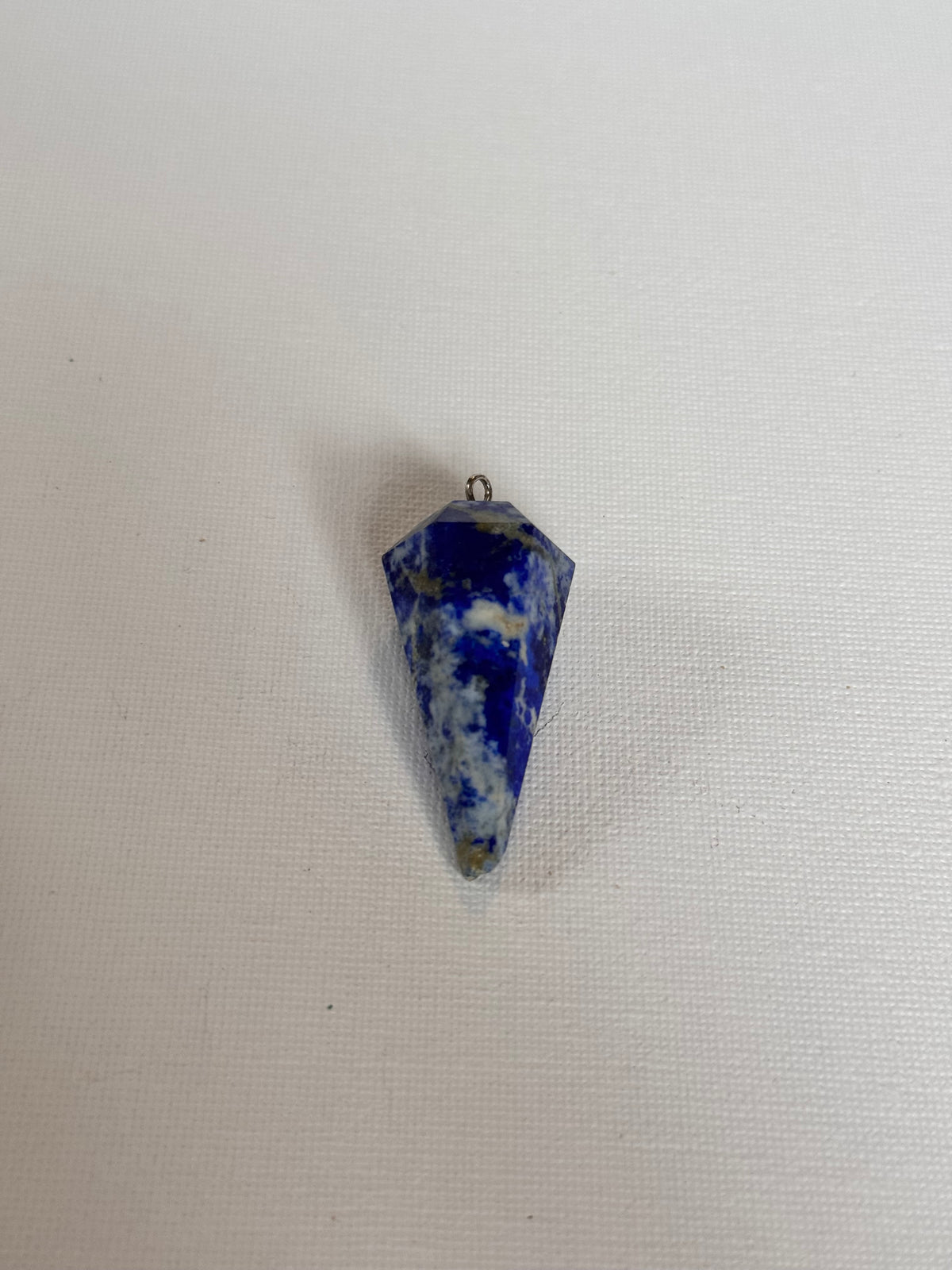 A small pointed blue and white marbled stone pendant or charm with a tiny metal loop at one end, placed on a plain light fabric background.