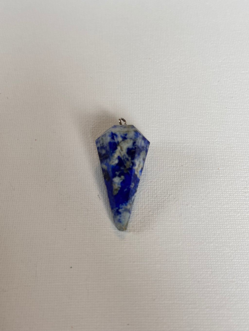 A small pointed blue and white marbled stone pendant or charm with a tiny metal loop at the top, placed on a white textured fabric background.