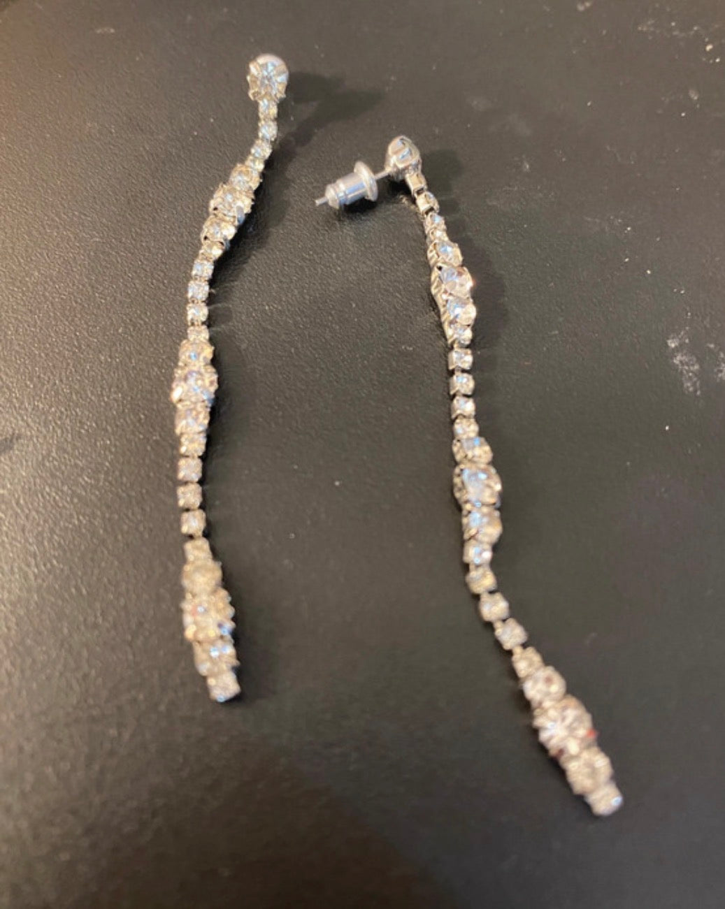 A pair of long, dangling statement earrings with clear rhinestones set in silver-toned metal, displayed on a dark textured surface.