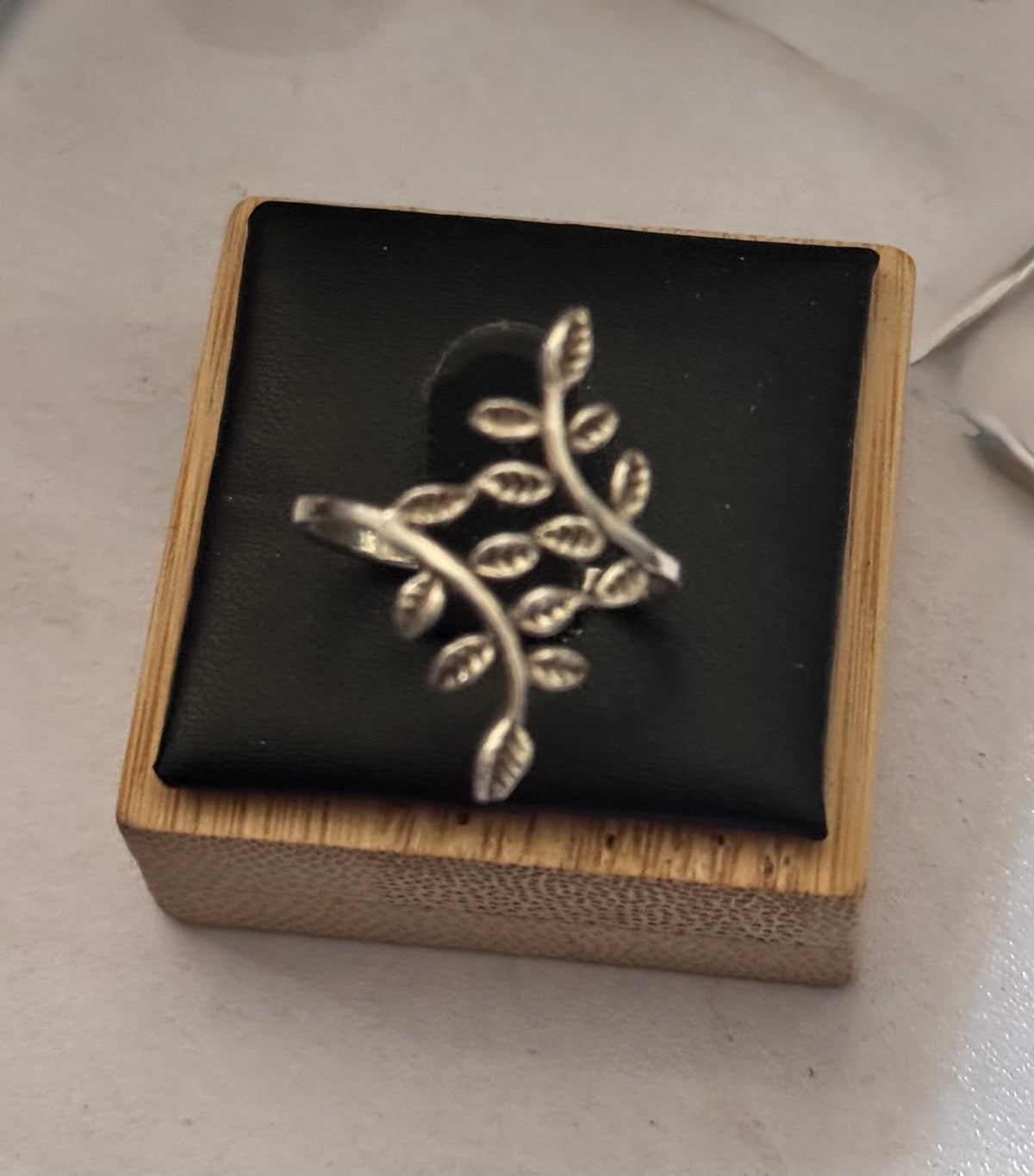 A close up of the sterling silver leaf-shaped ring displayed on a black cushion inside a small wooden box, placed on a light surface.