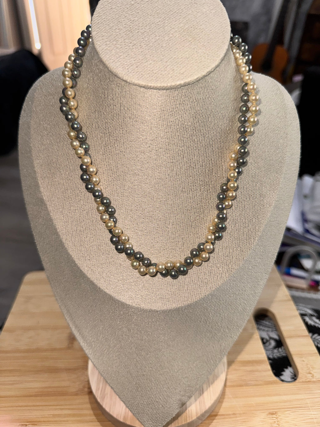 A double-strand beaded necklace with alternating dark gray and light beige pearls, displayed on a beige fabric jewelry bust.