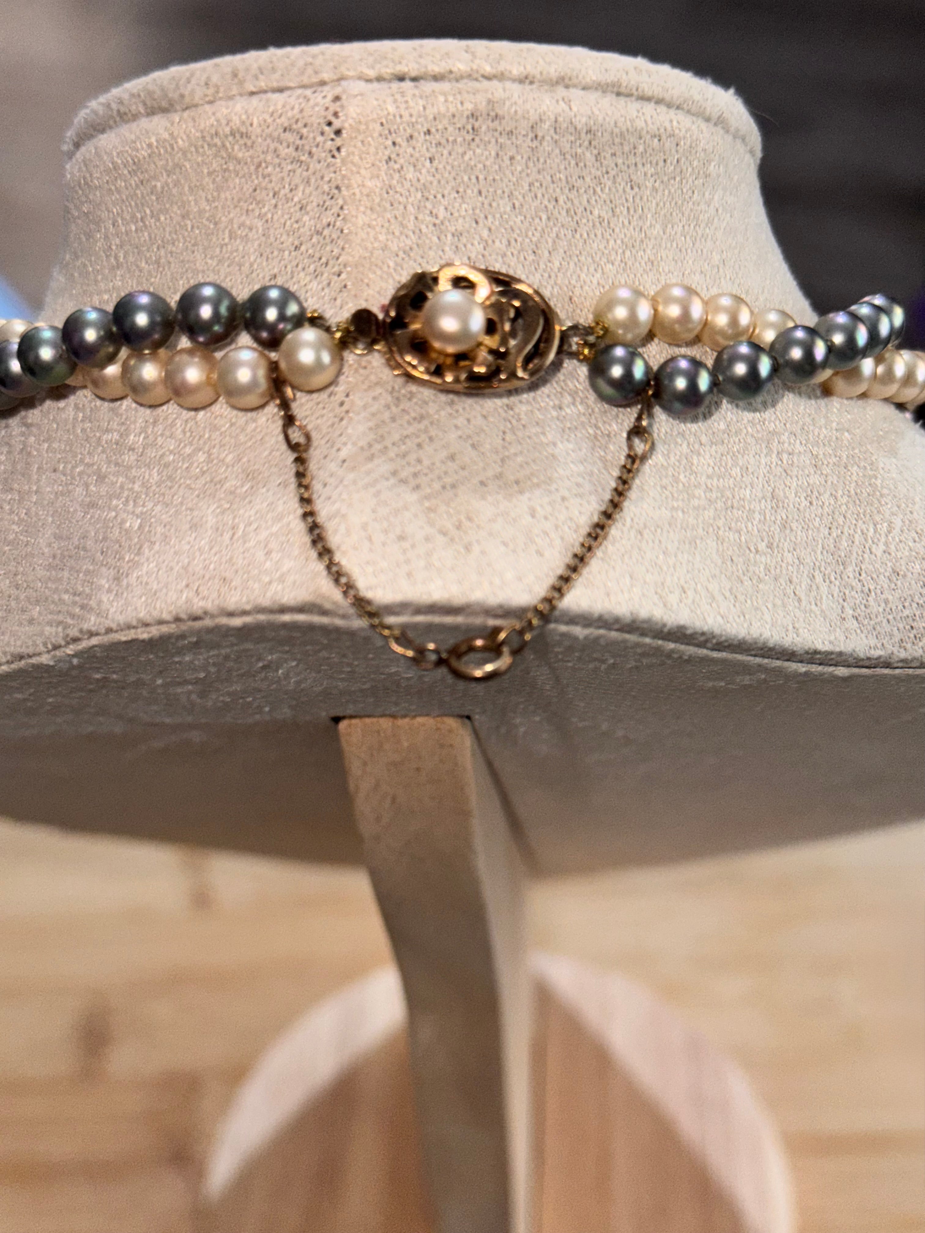 A double-strand pearl necklace with dark grey and cream pearls, featuring a bronze / gold-tone oval clasp with a central pearl and a delicate dangling gold chain, displayed on a beige fabric jewelry bust.