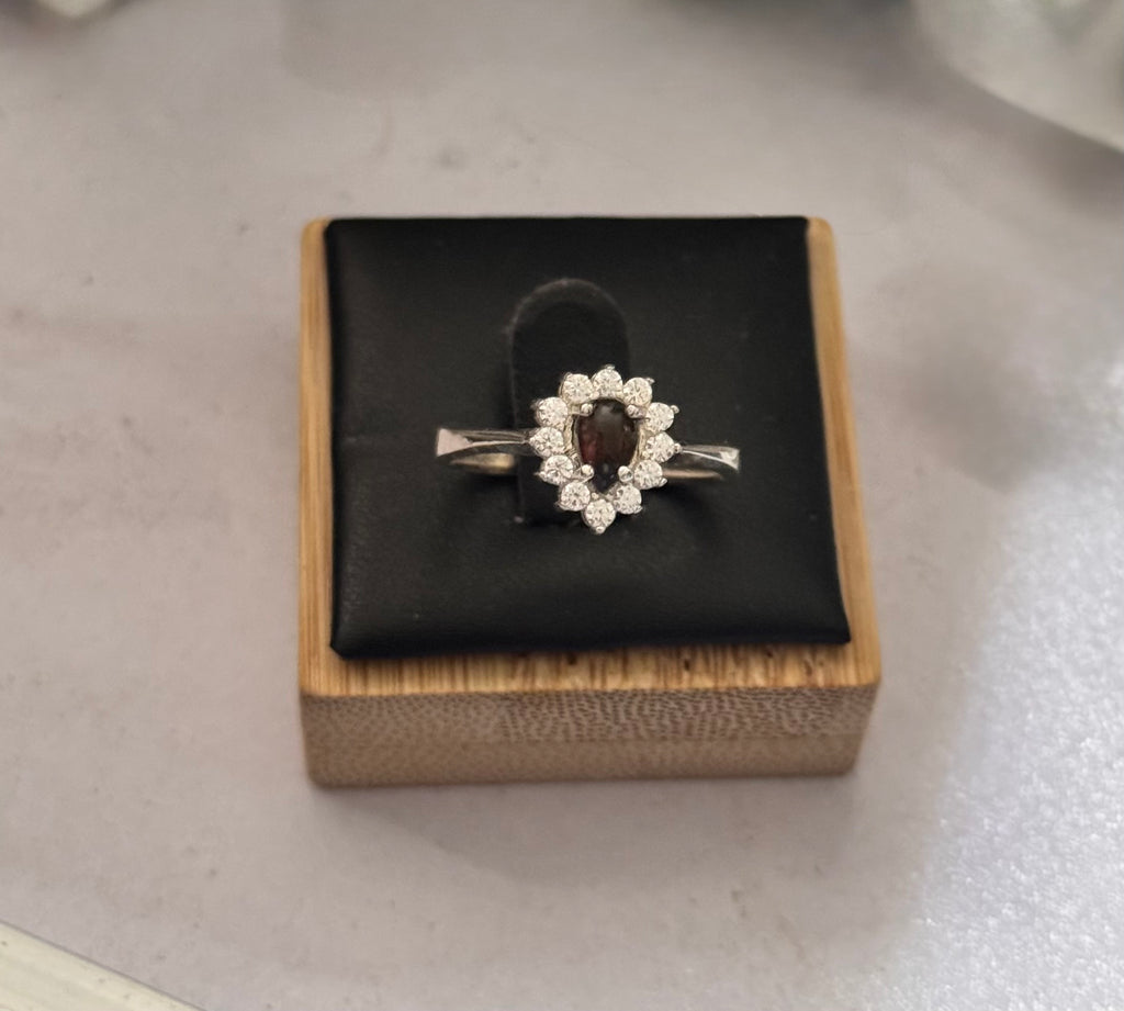 A sterling silver ring with a central dark red/green pearl gemstone surrounded by small clear stones, displayed on a black cushioned wooden box.
