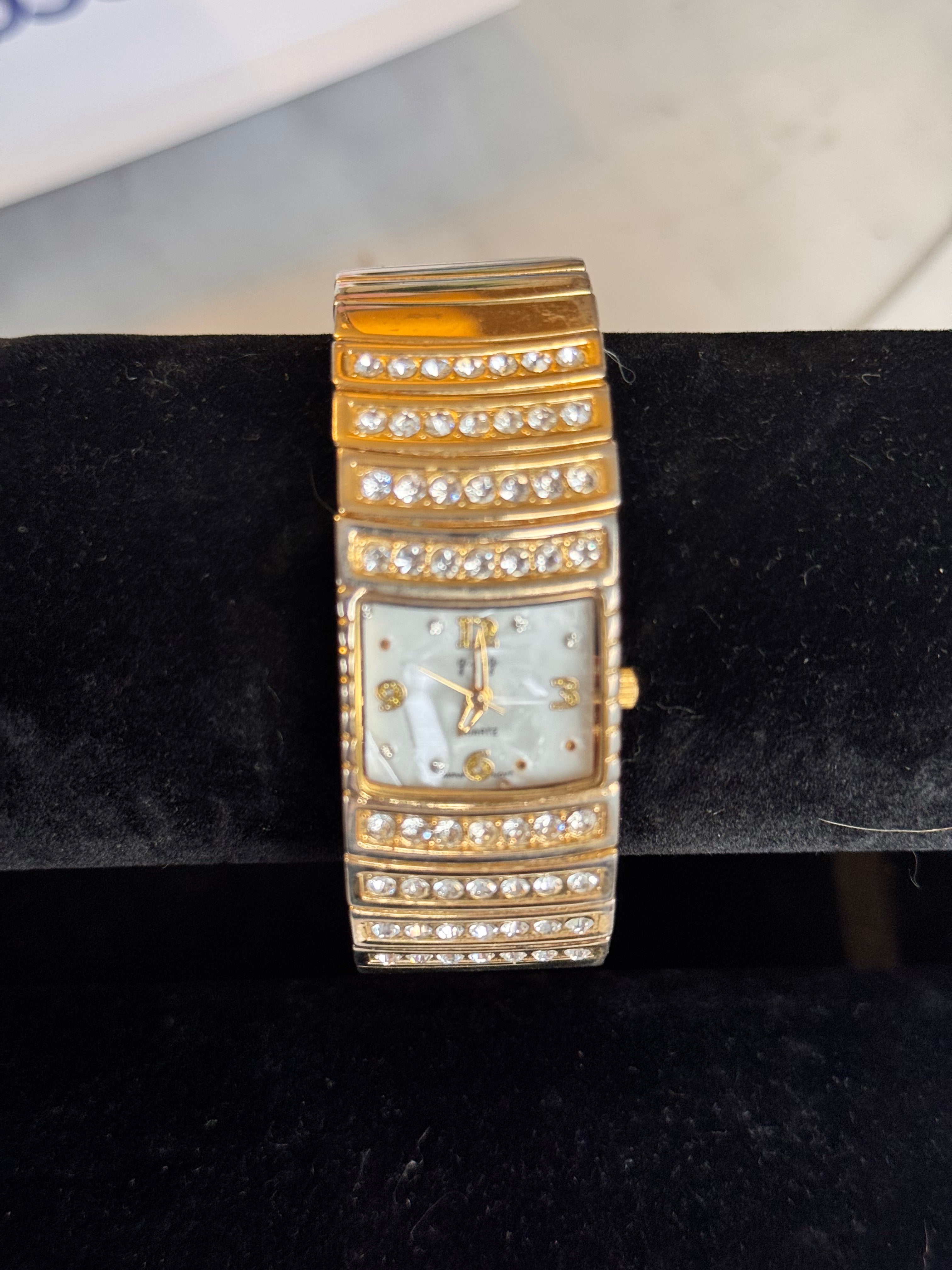 A gold-tone rectangular watch with a mother-of-pearl dial and rhinestone embellishments, displayed on a black velvet watch stand.