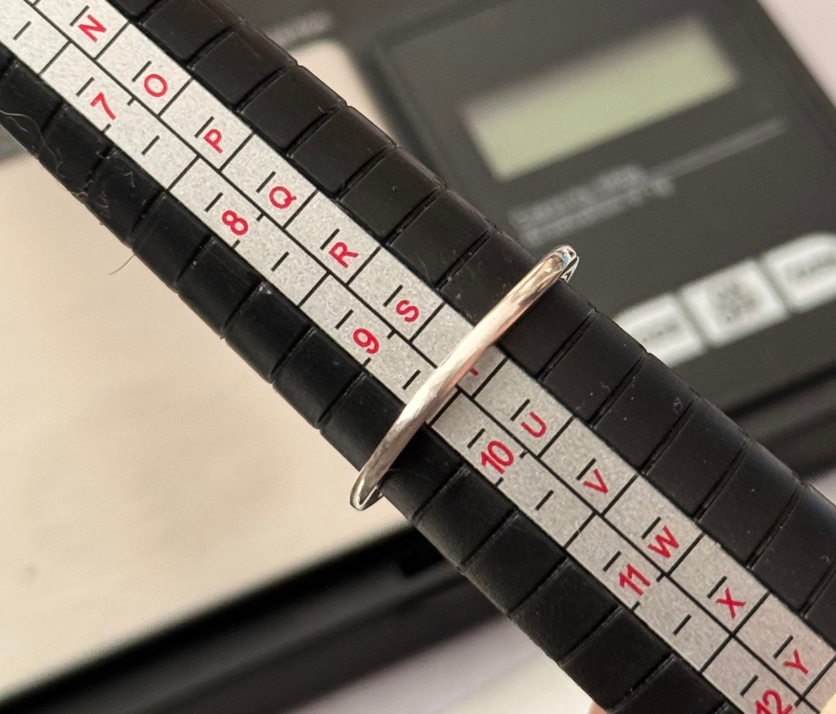 A silver ring displayed on a black ring sizer Size T with a white measurement scale, with a digital scale blurred in the background.
