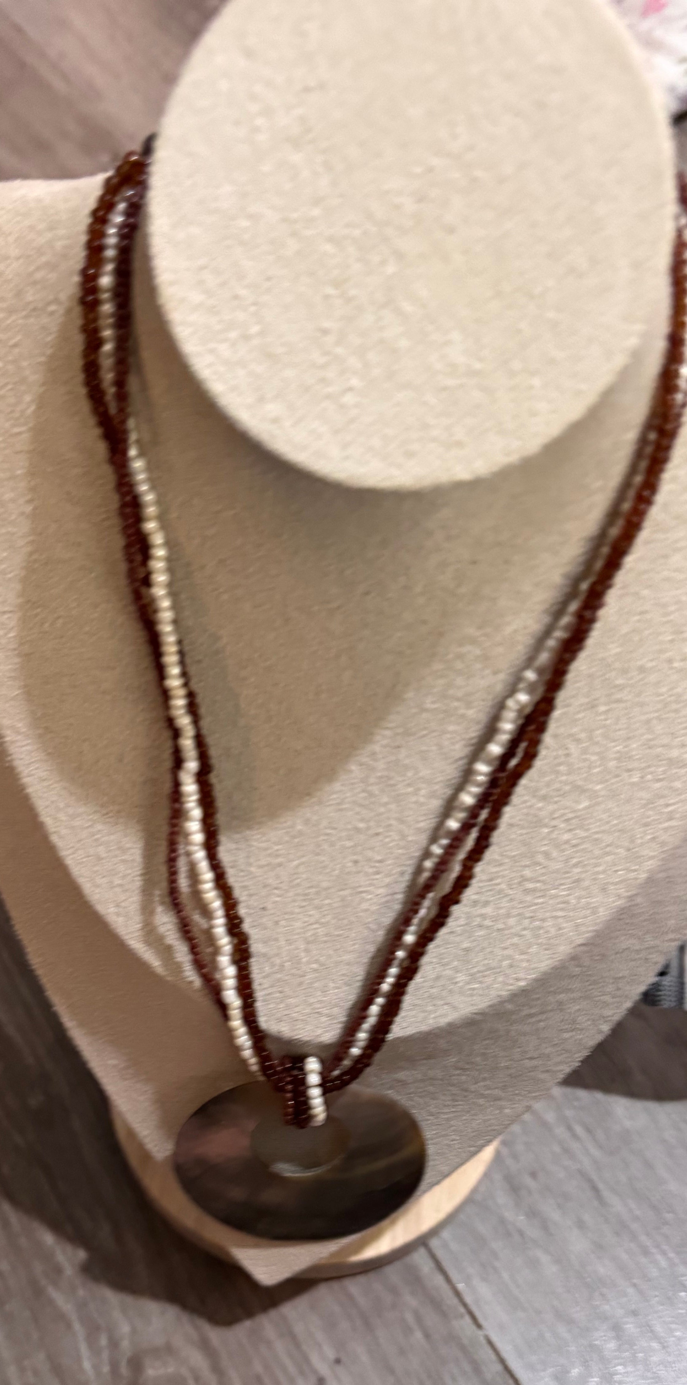 A beaded necklace with brown and cream strands and a large oval pendant, displayed on a beige jewelry bust. The bust is placed on a gray wood-look floor.