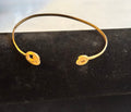 A thin gold cuff bangle with two small coils alternative heart charms, displayed on a black velvet jewelry stand.