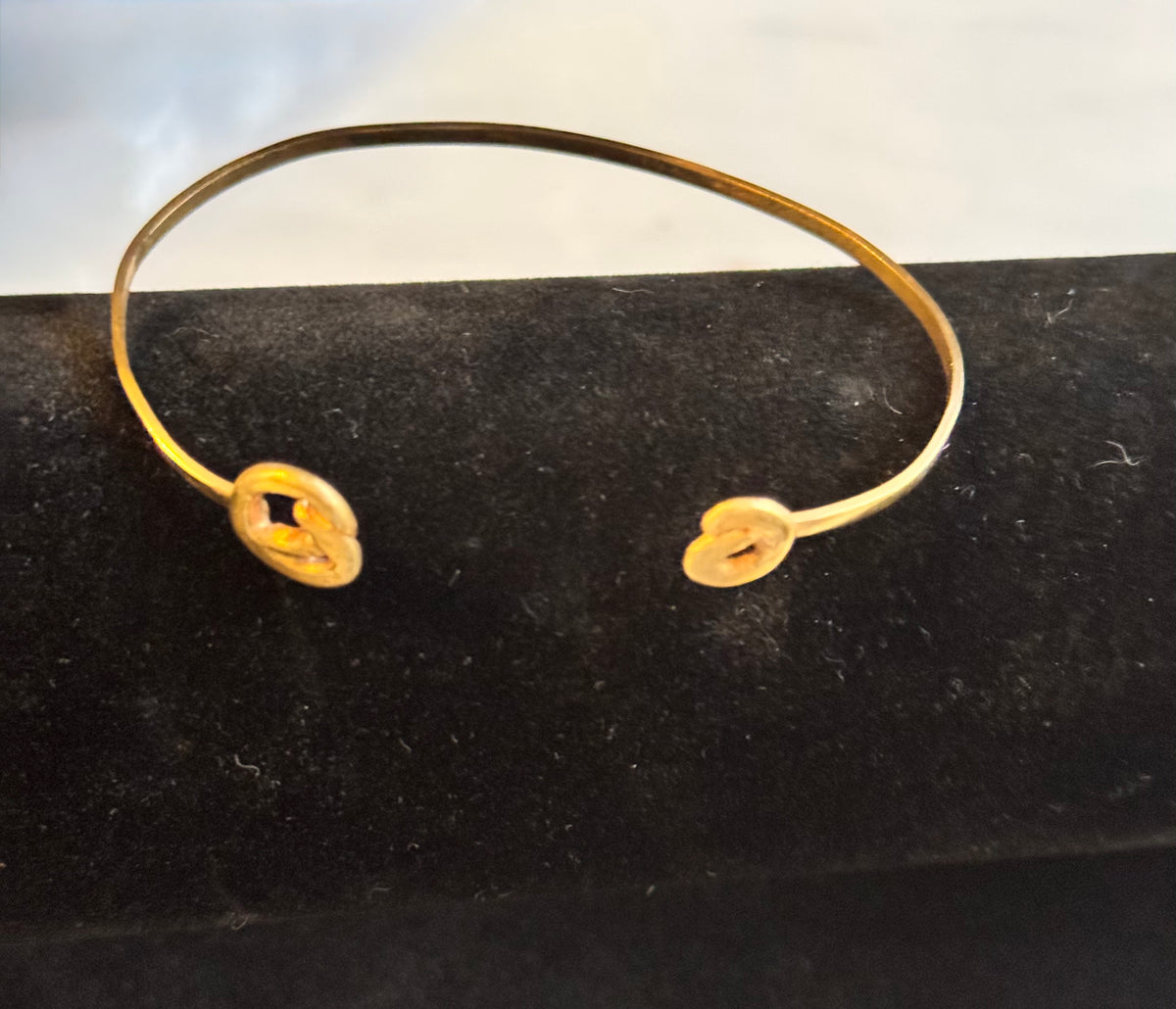 A thin gold cuff bangle with two small coils alternative heart charms, displayed on a black velvet jewelry stand.