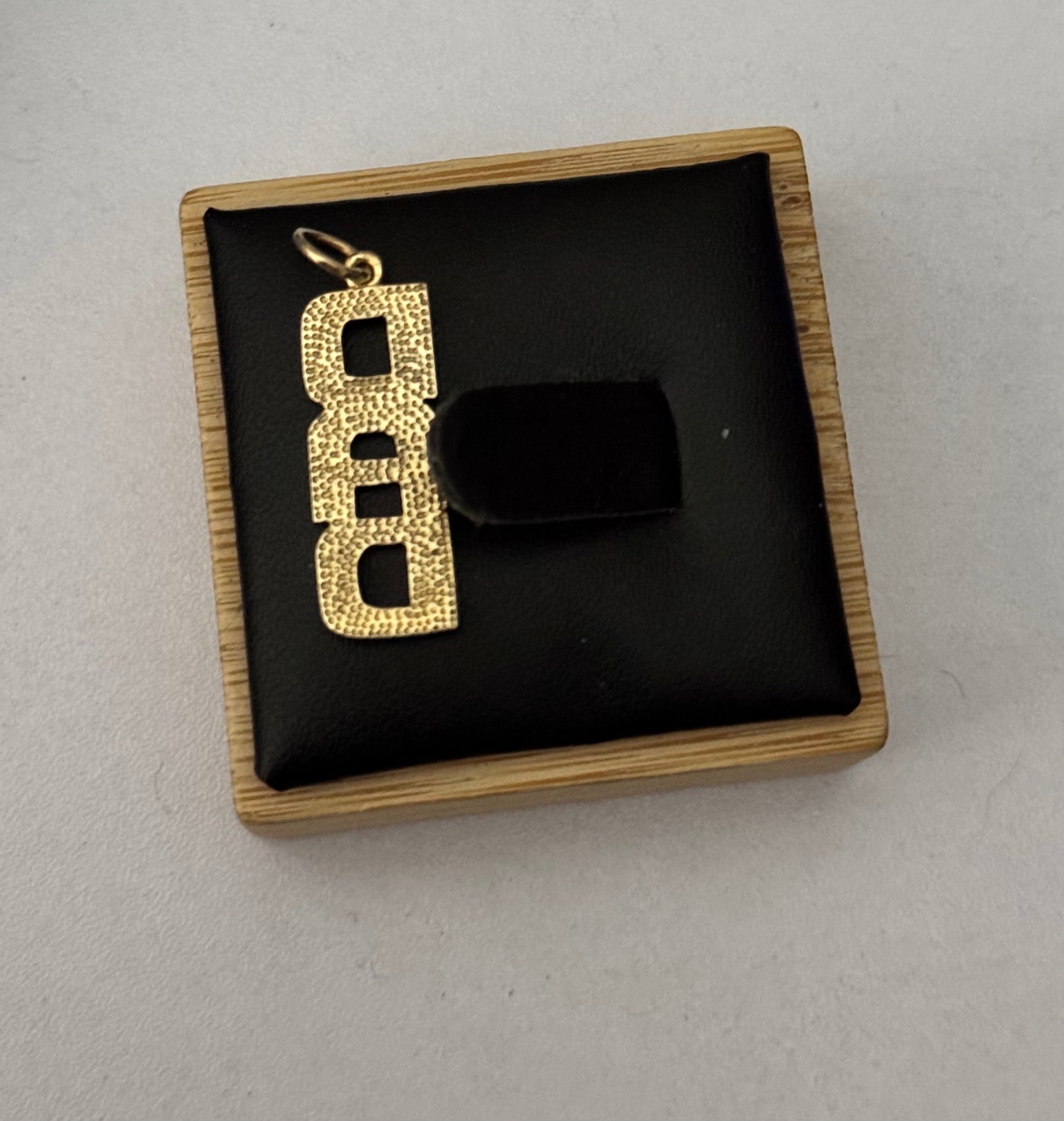 A 9ct hallmarked gold pendant with a textured, dotted pattern and a small top loop, displayed inside a square wooden jewelry box with a black cushion. The pendant is positioned next to a black velvet tag on a light gray background.