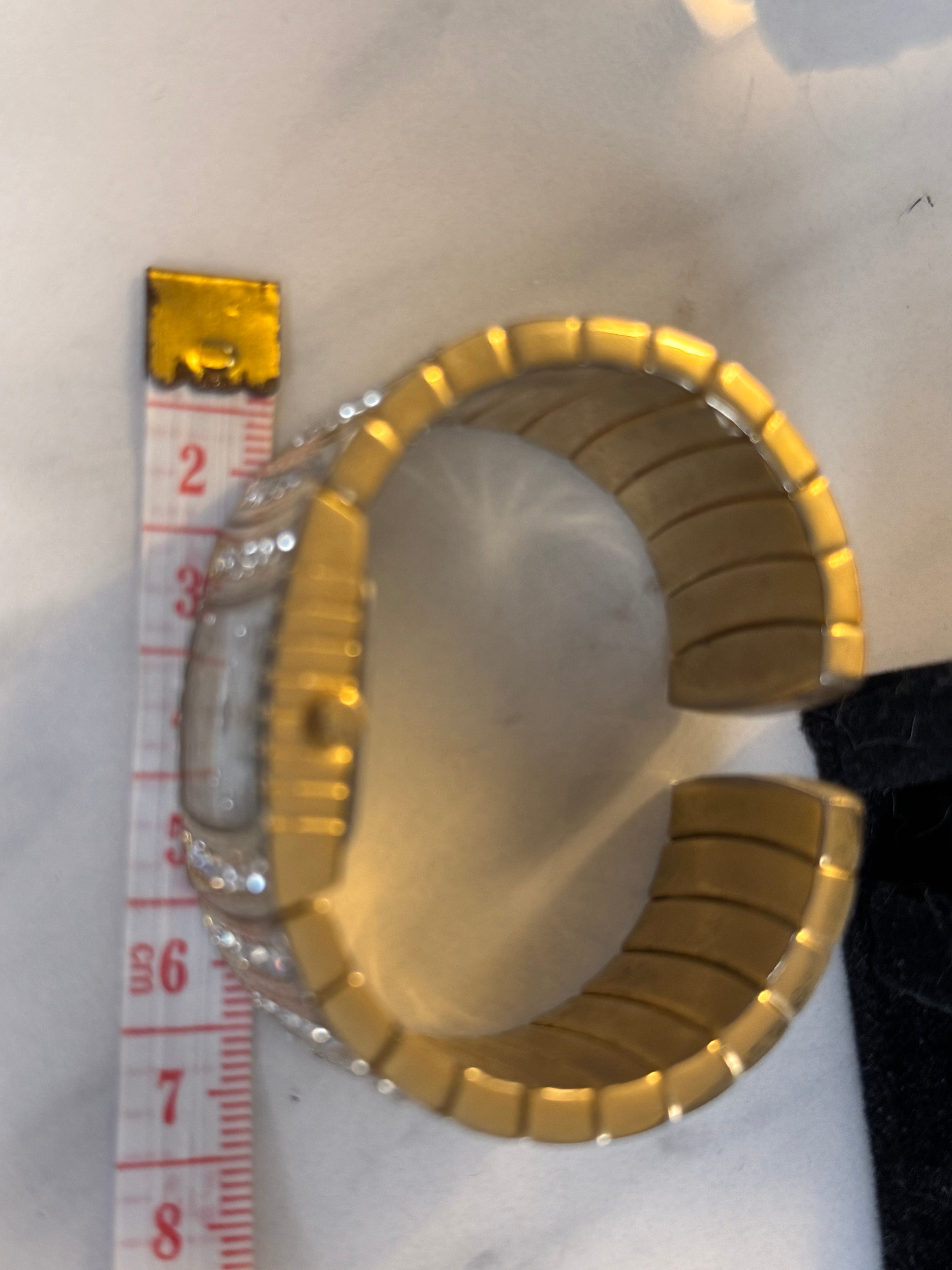 A gold-toned segmented cuff bracelet watch by GwG with clear rhinestone embellishments, shown next to a measuring tape to indicate scale on a light surface.