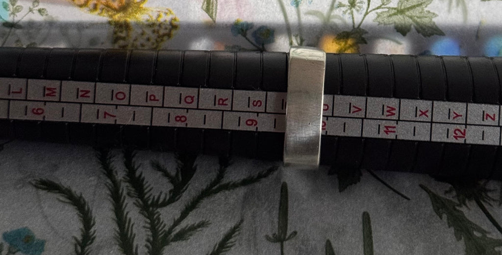 A sterling silver metal ring band shown on a black cylindrical measuring device with red and white size markings, placed on a floral fabric surface, to show the ring size of T.5