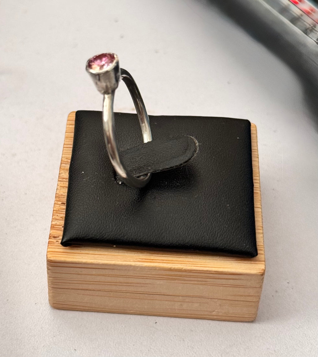 A silver ring with a 5mm pink moissanite gemstone displayed on a black cushion atop a light wood block stand.