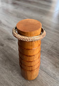 A gold-toned rhinestone bracelet displayed on a cylindrical wooden mandrel, stand, set against a grey wood-look floor background.