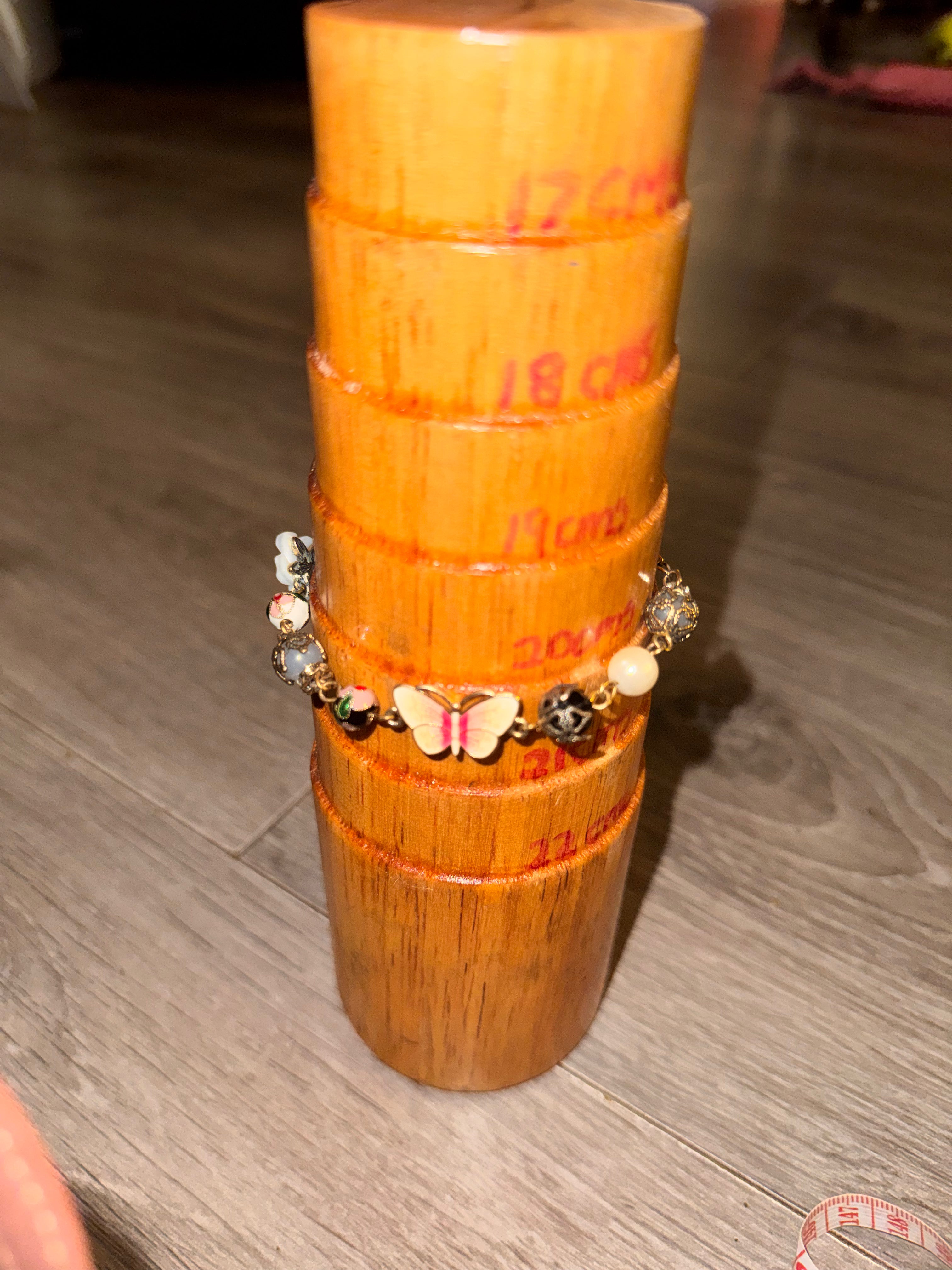A cylindrical wooden jewellery mandrel display on a grey wood floor, with a beaded charm bracelet featuring a butterfly enamel charm showing the measurements of the bracelet. 20-21cm 