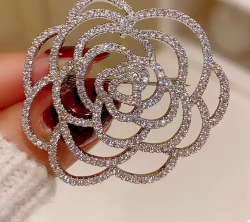 A close-up of a large, intricate rose-shaped brooch embellished with sparkling clear crystals on a silver-toned metal base, held by a hand with dark red nail polish.