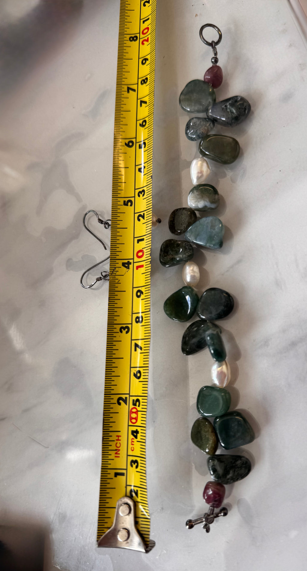A handmade beaded necklace with polished green and gray stones, white pearls, and red maroon beads, shown next to a yellow tape measure on a white marble surface. its an 8 inch bracelet