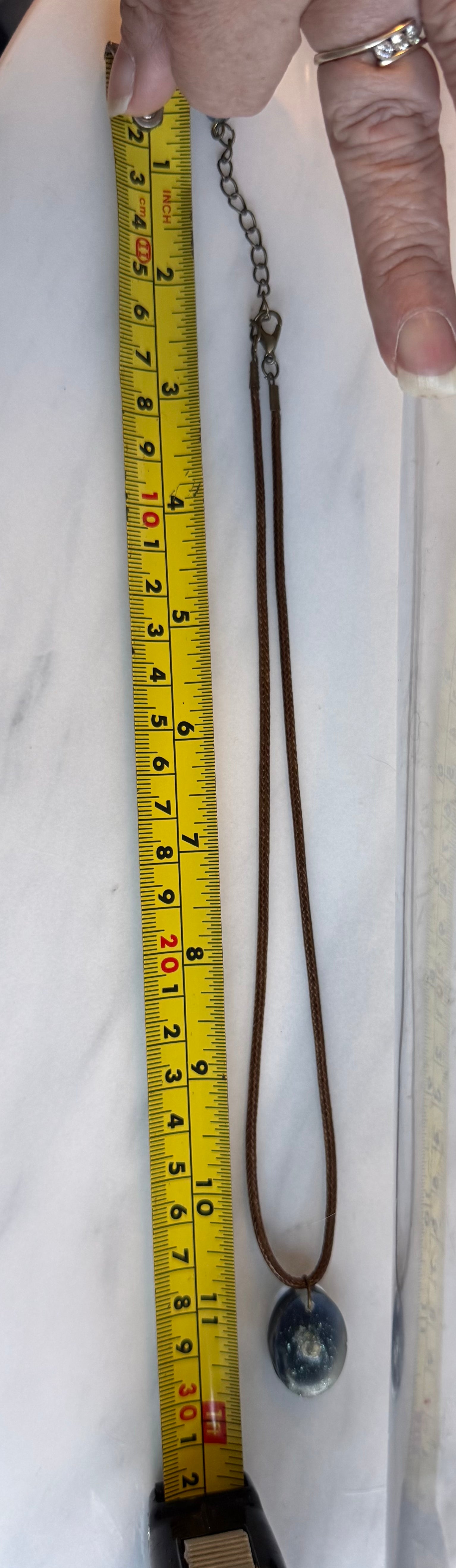 A brown leather cord necklace with a round dark blue-gray stone pendant, shown next to a yellow tape measure for size reference. A hand is holding the top of the necklace. From the extensor to the tip of the pendant with the necklace closed is 30cm