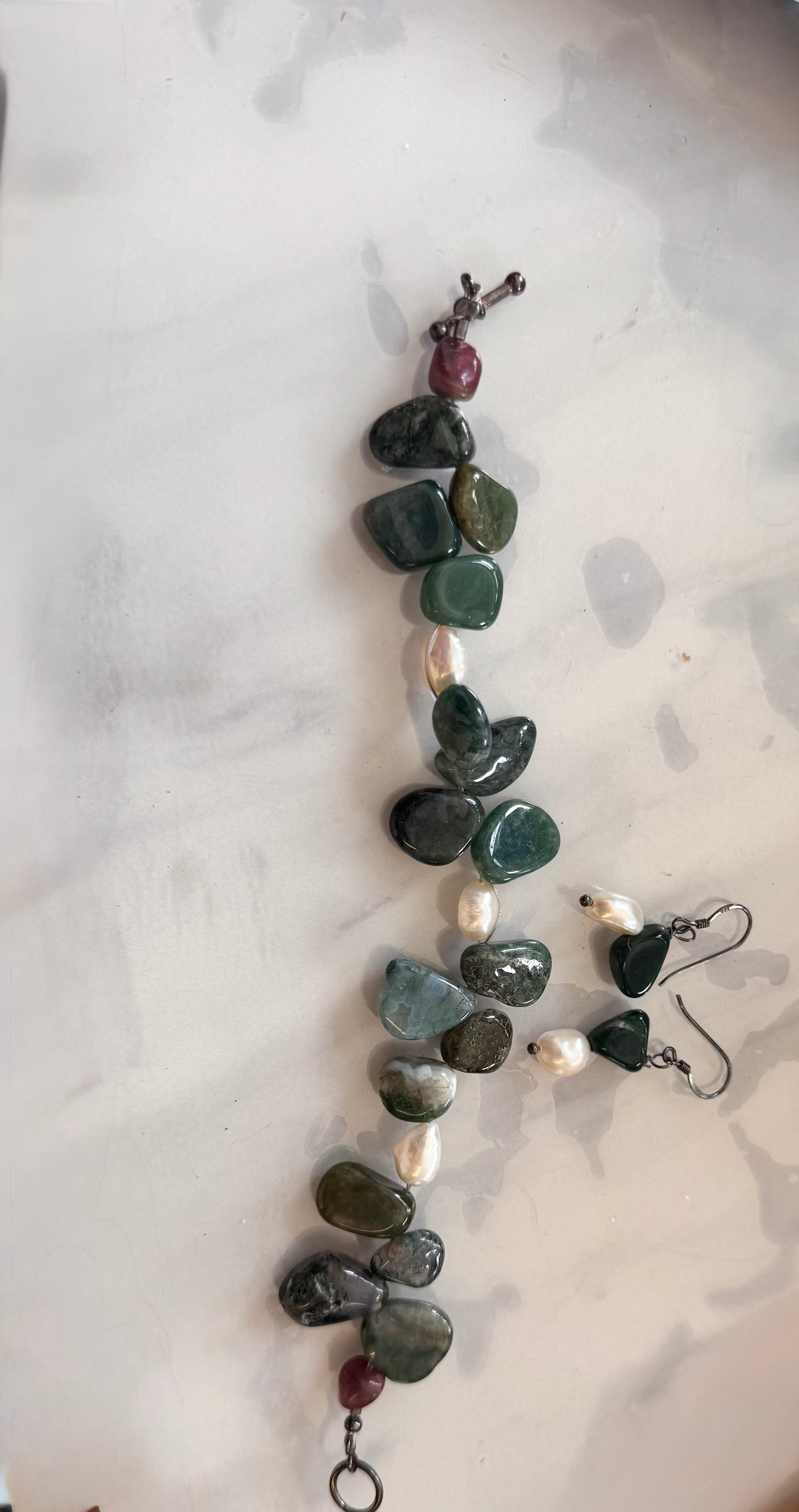 A handmade beaded bracelet and matching dangle earrings made with polished dark green stones (moss agate), white pearl beads, and small red accent beads, displayed on a light marble surface.