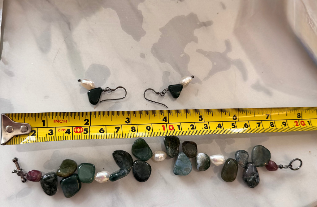 A pair of small drop earrings and a matching beaded bracelet laid out on a white marbled surface with a yellow tape measure in front for scale. The jewelry appears to use dark green and gray polished stones with white pearl-like beads and small red accent beads, attached to silver tarnished earring hooks and clasps.
