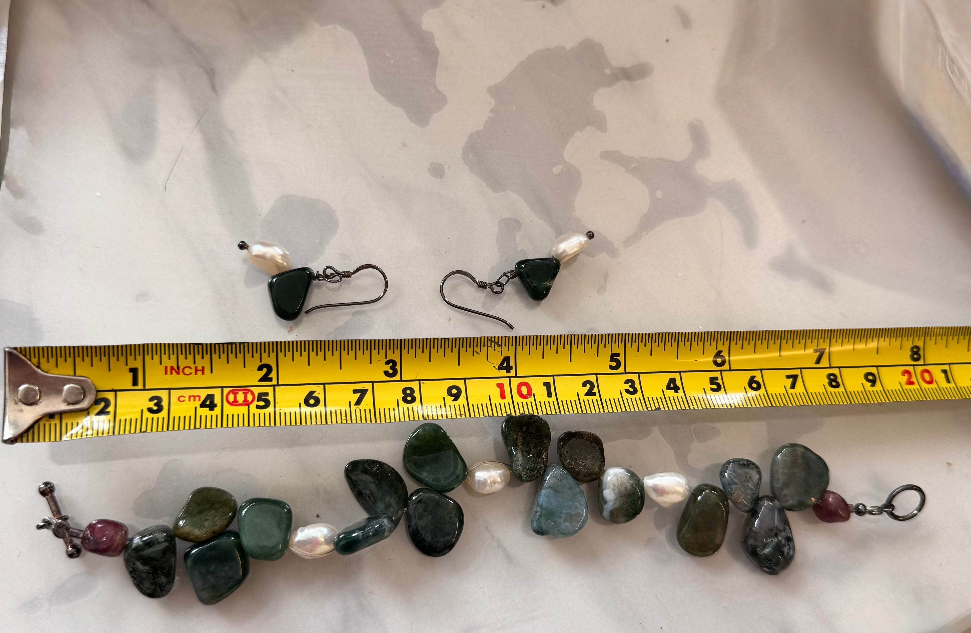 A pair of small drop earrings and a matching beaded bracelet laid out on a white marbled surface with a yellow tape measure in front for scale. The jewelry appears to use dark green and gray polished stones with white pearl-like beads and small red accent beads, attached to silver tarnished earring hooks and clasps.