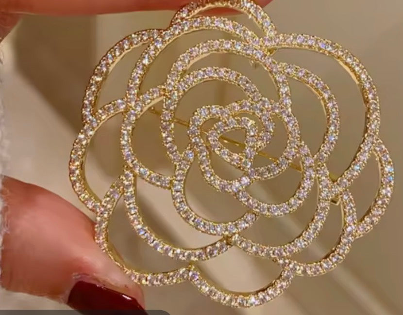 A gold-tone rose-shaped brooch or pin embellished with clear pavé-style crystals, held between fingers against a soft beige background.