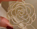 A gold-tone rose-shaped brooch or pin embellished with clear pavé-style crystals, held between fingers against a soft beige background.