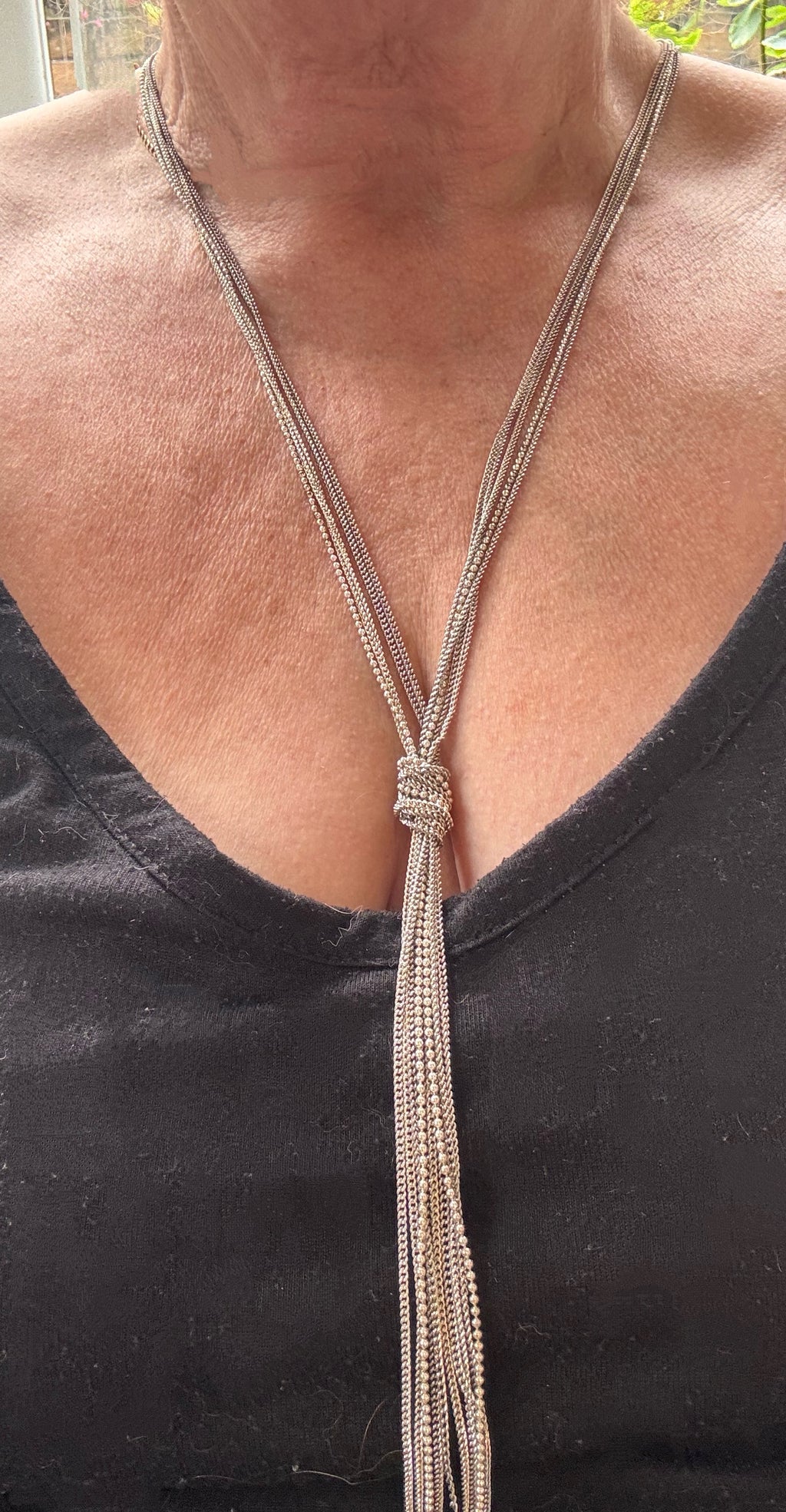 A close-up of a person wearing a long silver-toned lariat-style necklace with a knotted center and long dangling strands, paired with a black V-neck top. This is a real image not AI