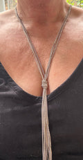 A close-up of a person wearing a long silver-toned lariat-style necklace with a knotted center and long dangling strands, paired with a black V-neck top. This is a real image not AI