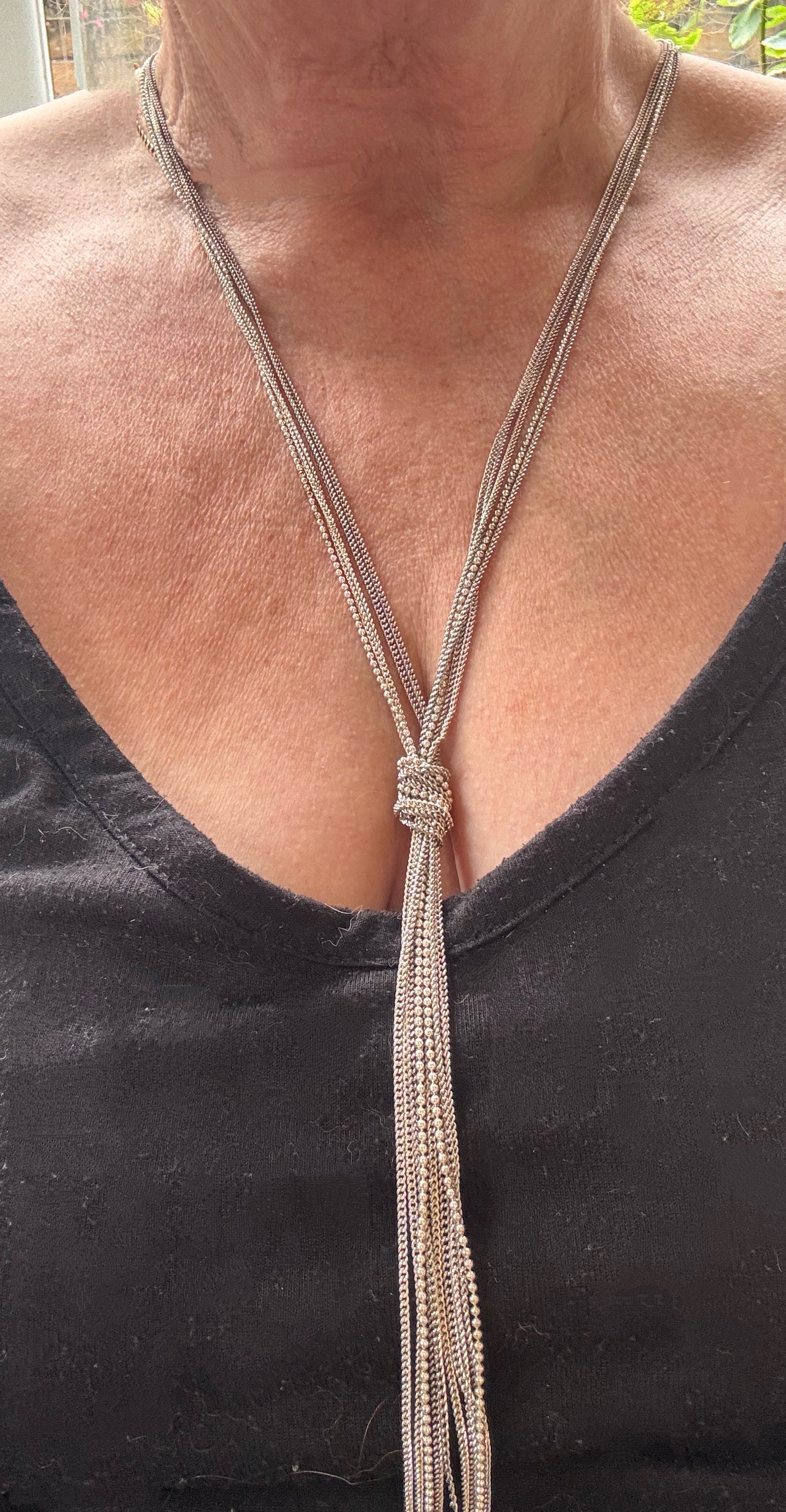 A close-up of a person wearing a long silver-toned lariat-style necklace with a knotted center and long dangling strands, paired with a black V-neck top. This is a real image not AI