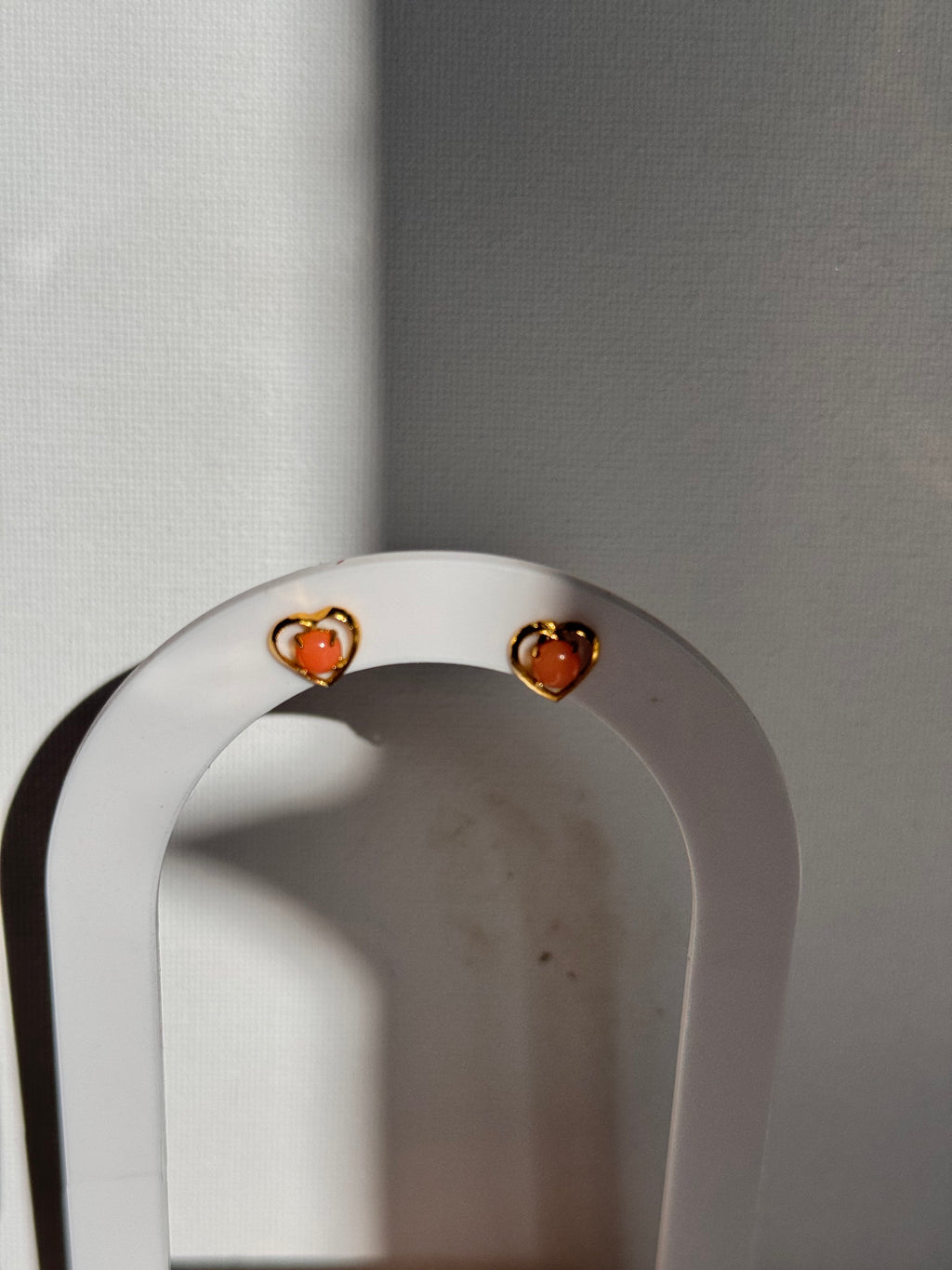 A close-up of a white curved jewelry display stand holding two small gold-toned earrings with peach-pink stones, set against a light gray background.