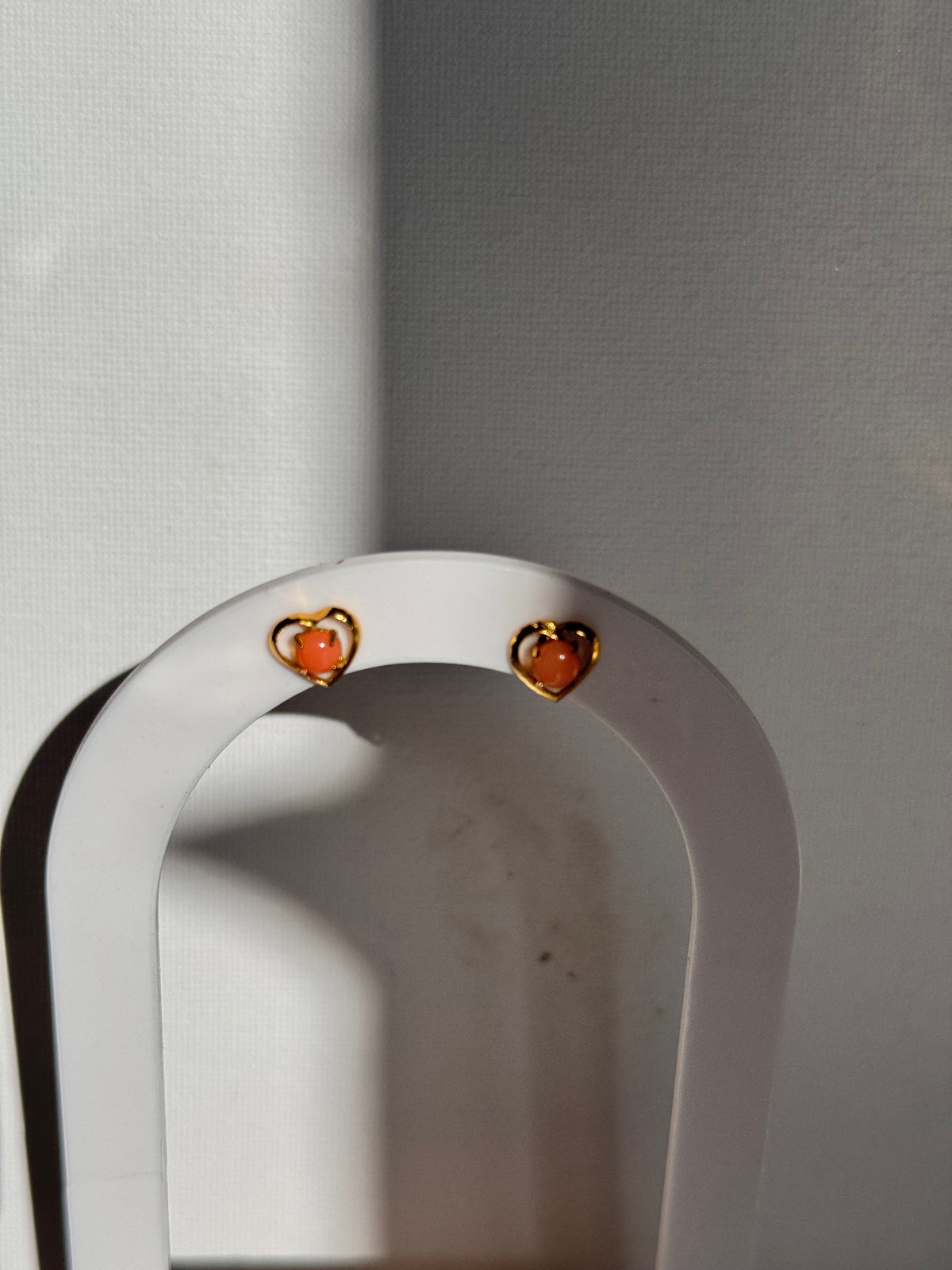 A close-up of a white curved jewelry display stand holding two small gold-toned earrings with peach-pink stones, set against a light gray background.