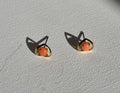 A pair of gold plated stud earrings with oval coral-pink gemstones, displayed on a textured white fabric surface with sharp shadows.