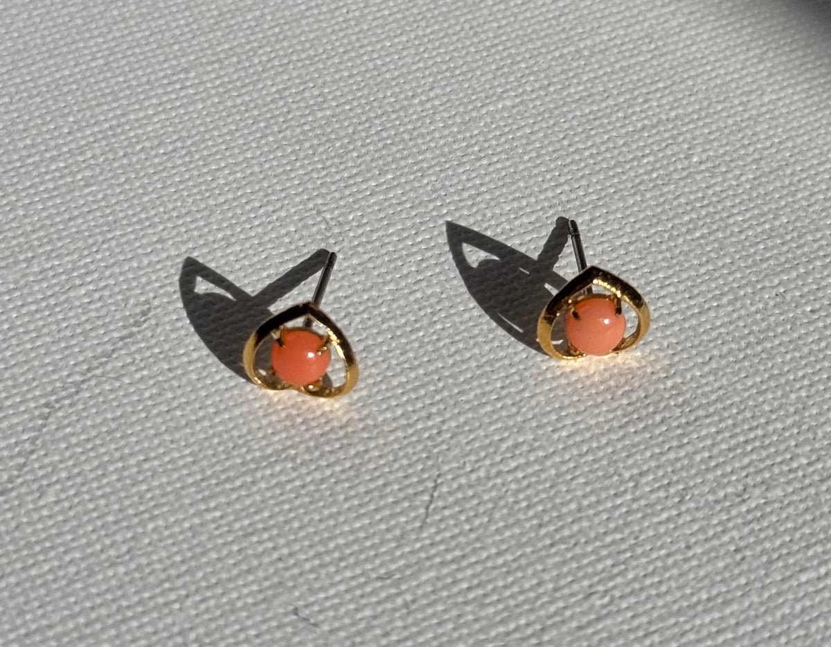 A pair of gold plated stud earrings with oval coral-pink gemstones, displayed on a textured white fabric surface with sharp shadows.