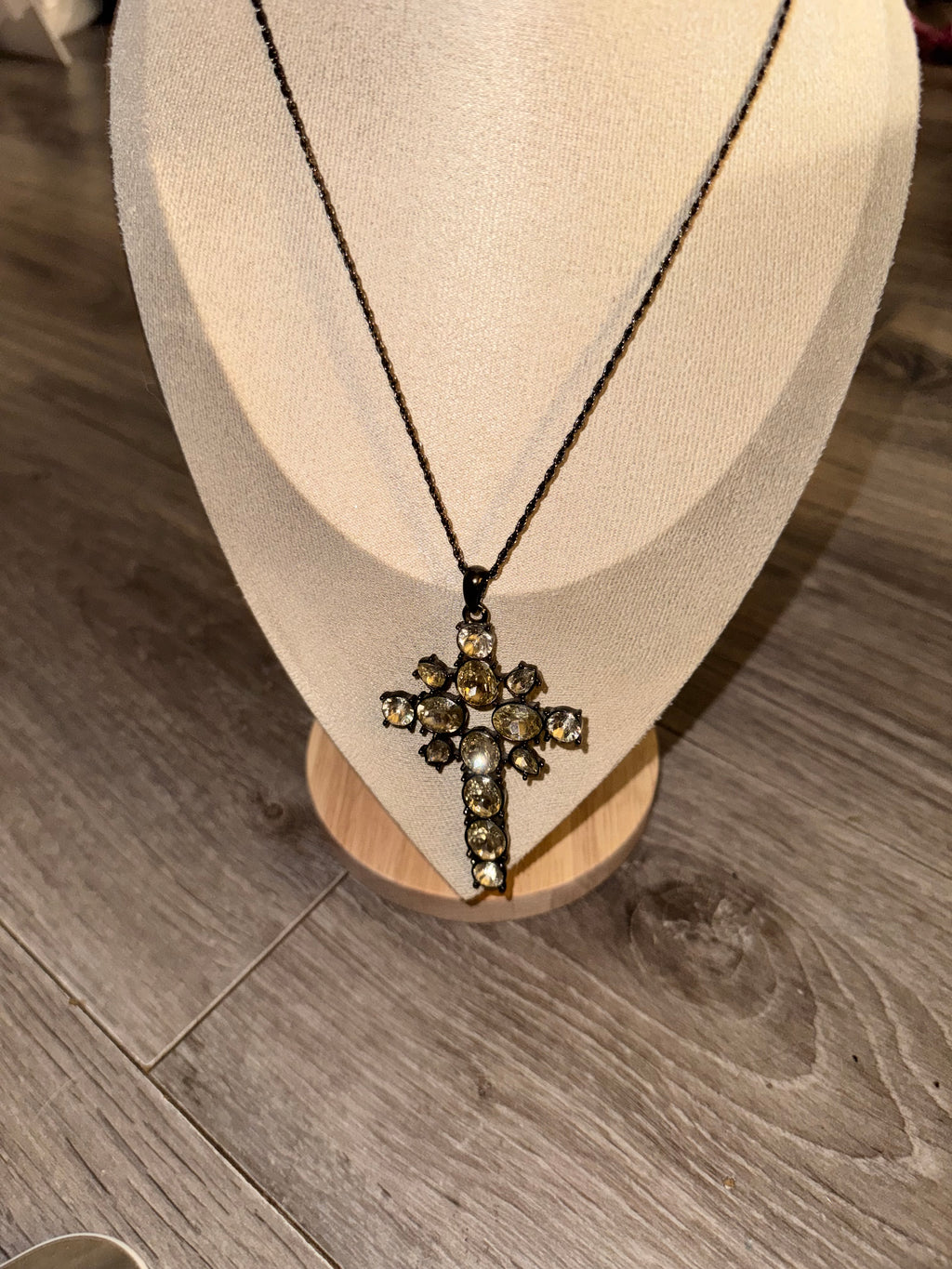 A close-up of a necklace displayed on a cream fabric jewelry bust, featuring a dark chain and a decorative Gothic Style Cross with clear stones and metallic details, set against a gray wood floor background.