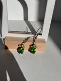 A pair of dangling earrings with green faceted rhinestones Emerald green colour and small metallic beads, hanging from silver-toned hooks on a wooden display block against a textured gray wall.