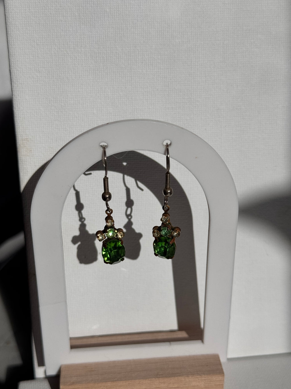 A pair of dangling earrings with green faceted rhinestones and metal settings, displayed on a white U-shaped jewelry stand.
