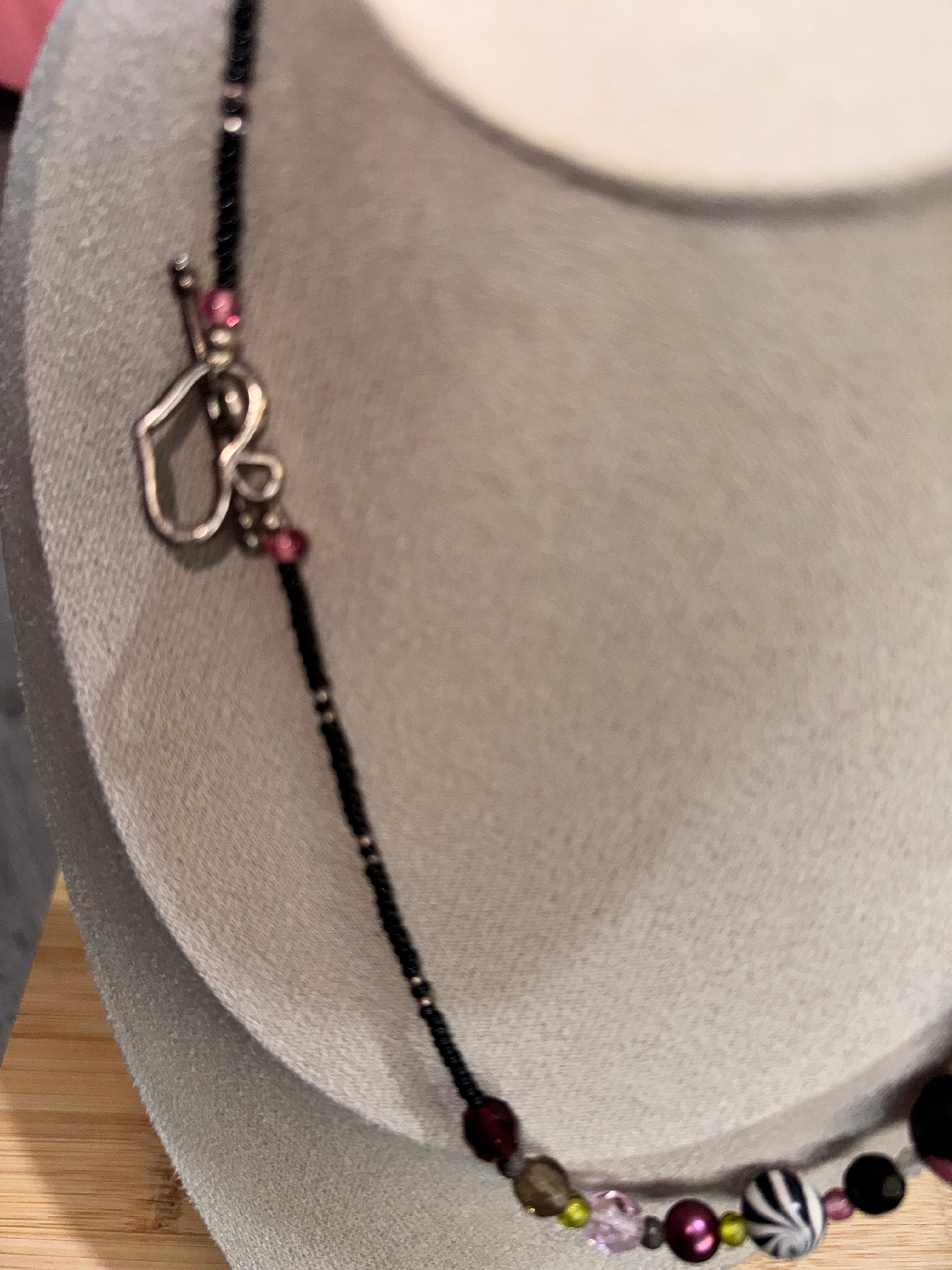 A beaded necklace with a silver-toned open heart clasp, displayed on a gray velvet jewelry bust. The necklace features a black beaded base with various colorful accent beads in pink, purple, green, and a black-and-white patterned bead.