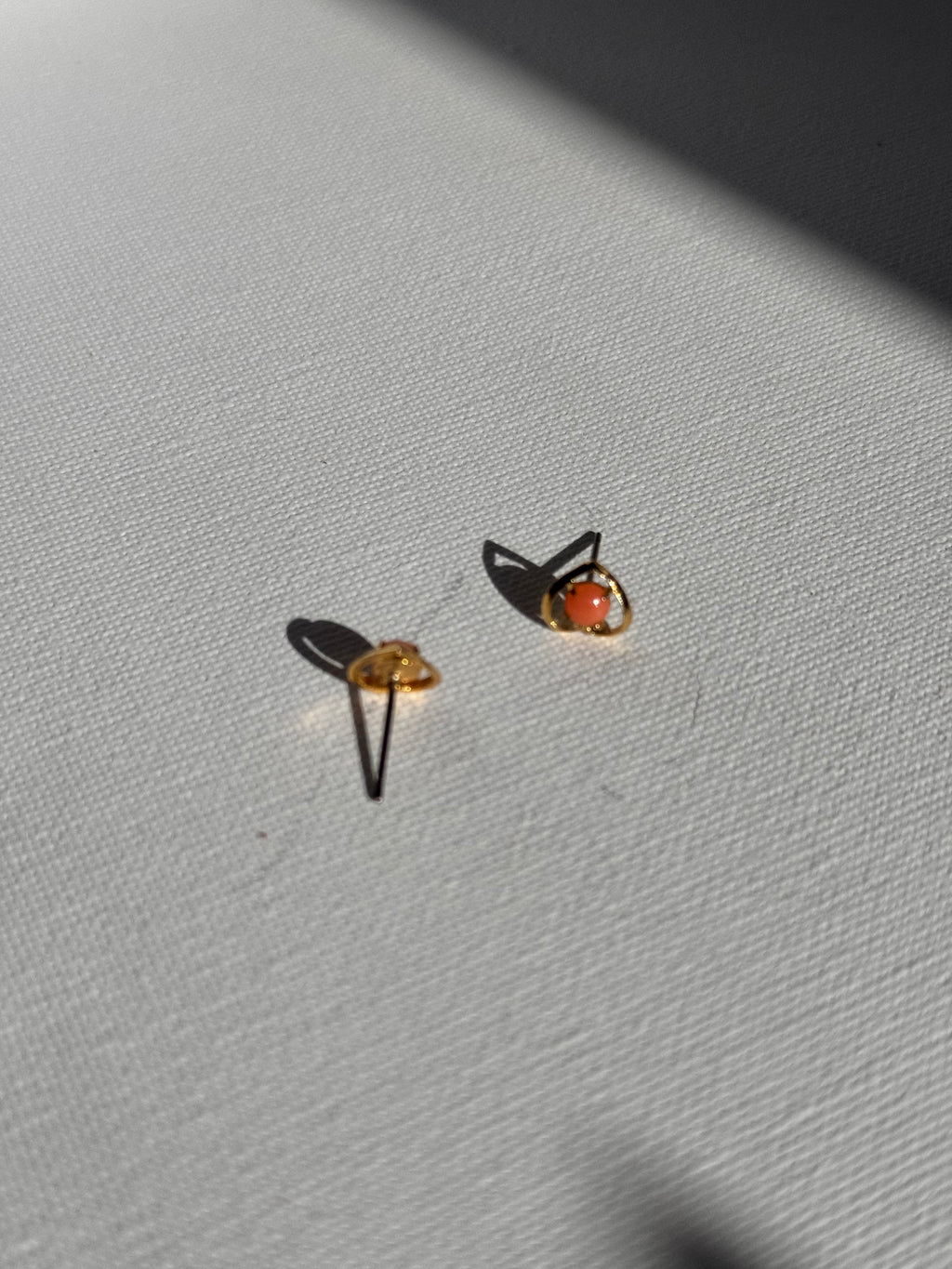 A pair of small gold plated heart stud & post earrings with round orange/coral-pink gemstones, displayed on a textured white surface under harsh direct sunlight.