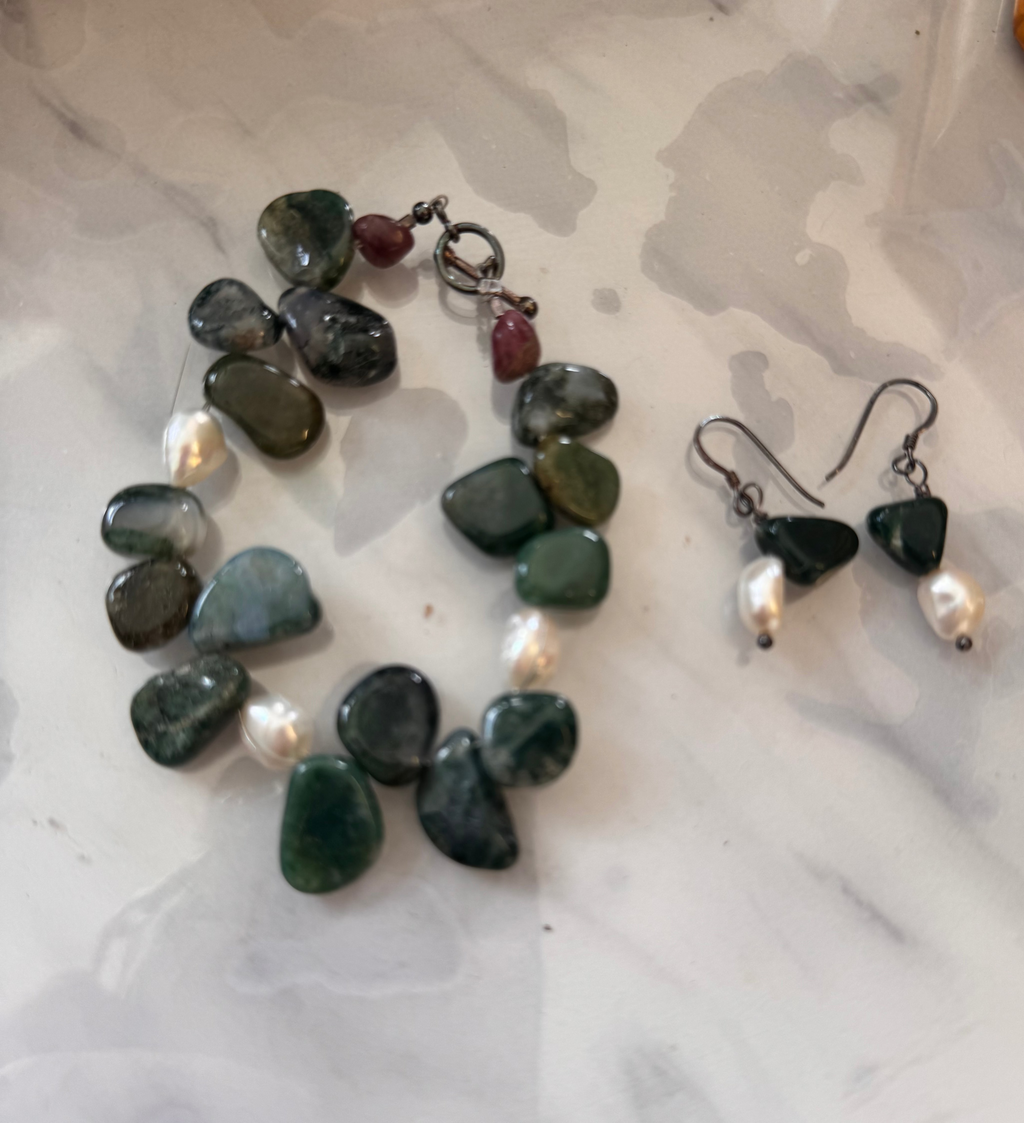A jewelry set featuring a tumbled stone bracelet and matching drop earrings on a white and gray marble surface. The stones are primarily dark green (moss agate) and gray with small white pearls and a tiny burgundy accent bead on the bracelet.