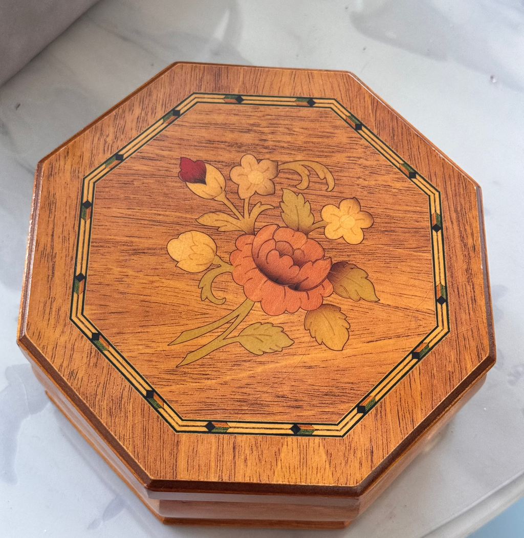 An octagonal wooden Jewellery box with a floral inlay design on the lid, featuring a central orange rose and yellow flowers, resting on a light marble surface.