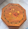 An octagonal wooden Jewellery box with a floral inlay design on the lid, featuring a central orange rose and yellow flowers, resting on a light marble surface.