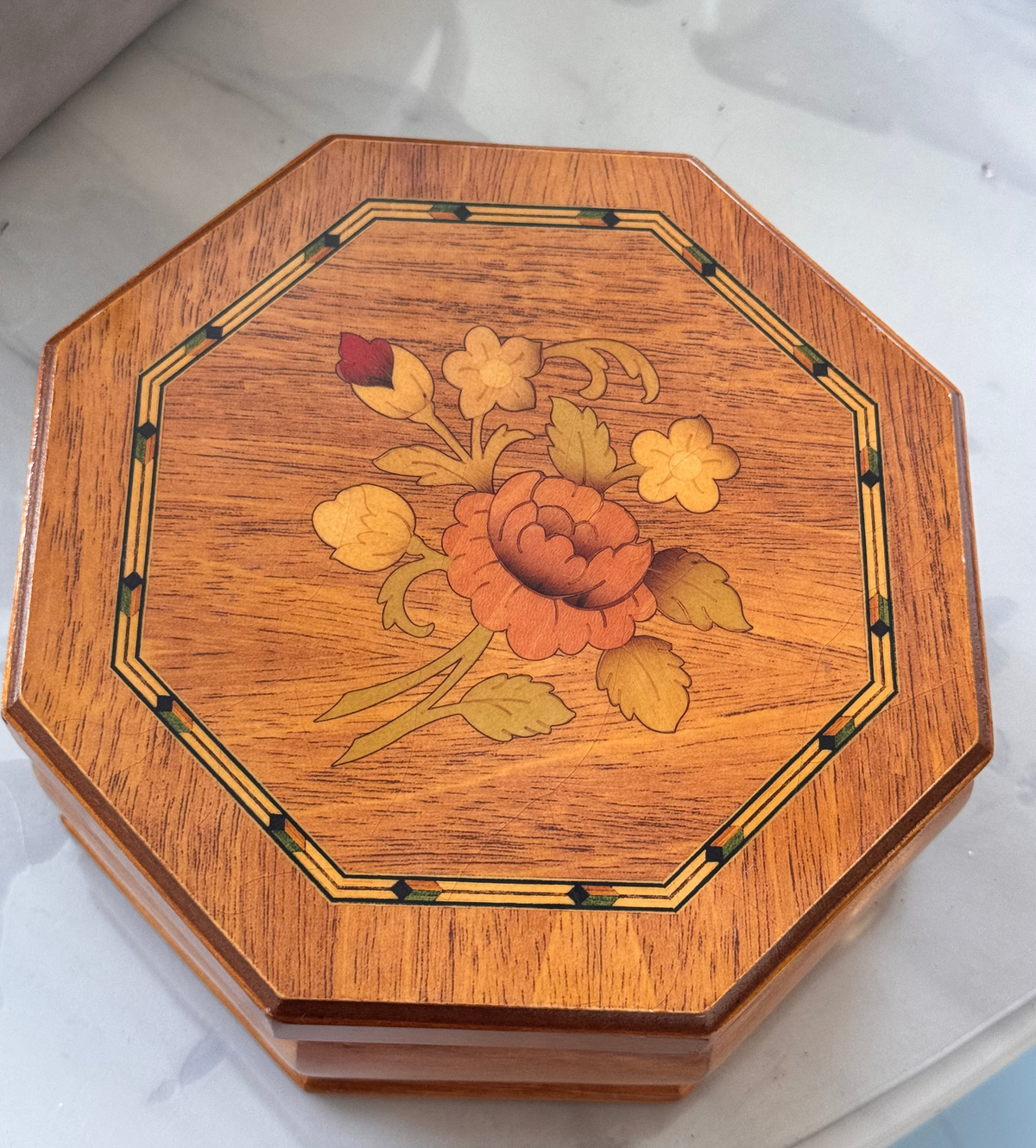 An octagonal wooden box with a floral inlay design on the lid, featuring orange, yellow, and red flowers with green leaves, set against a light marble background.