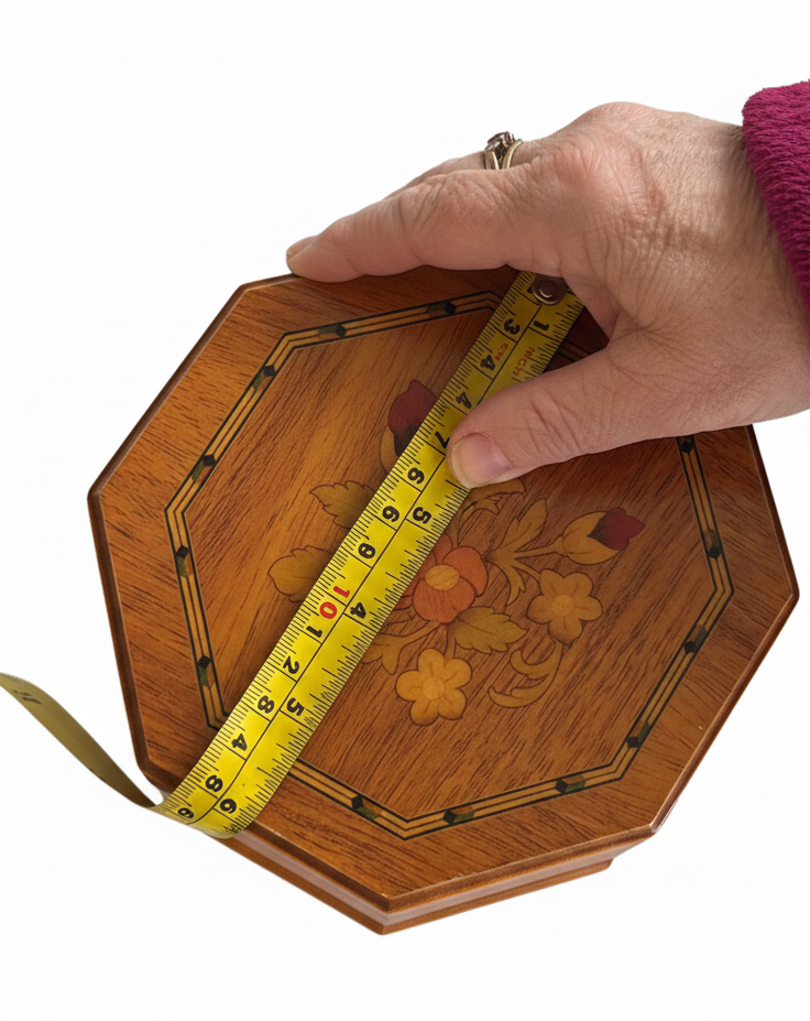 An octagonal wooden jewellery box with floral inlay and is shown with a hand holding a yellow tape measure across the top to indicate its size, on a plain white background. 6 x 6 inches
