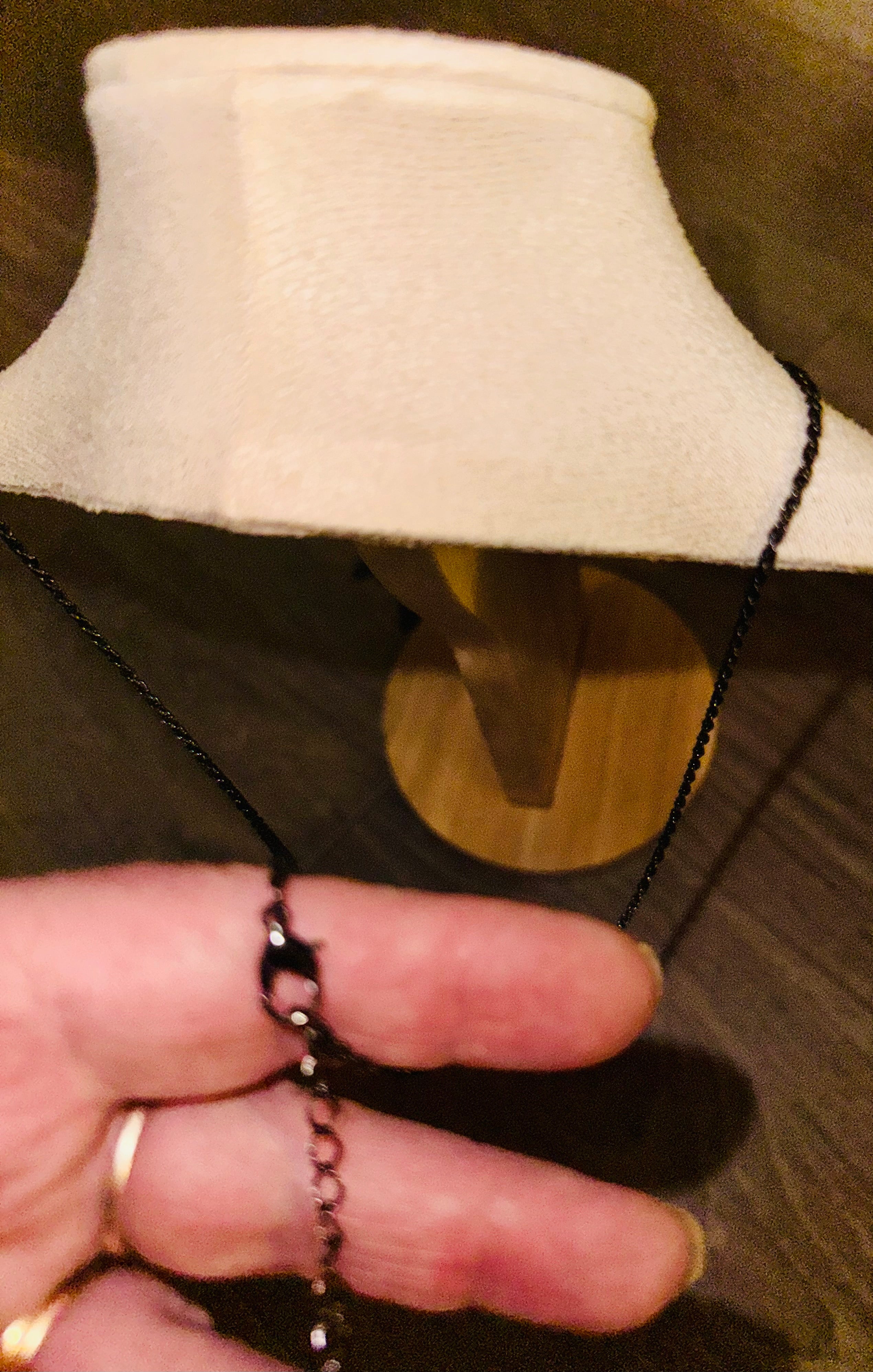 A close-up of a hand holding a black chain necklace with a dark metal clasp, with a cream-colored fabric jewelry display bust in the background.