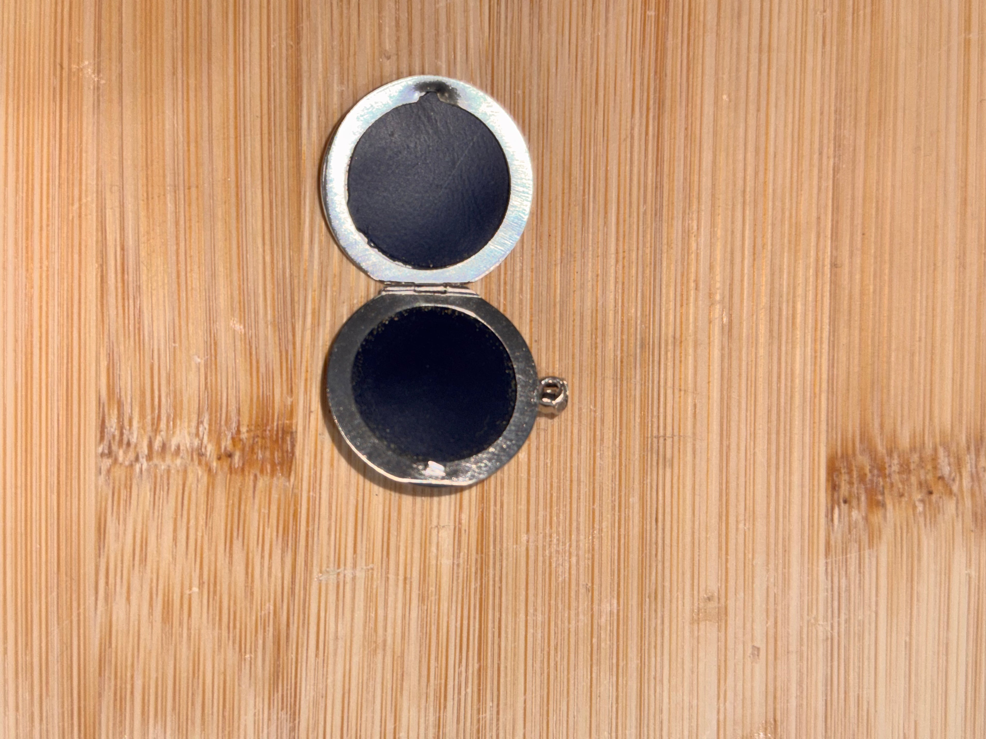 A small round silver metal locket or pendant with an open hinged lid, showing dark interior lining, placed on a light brown bamboo wood background.