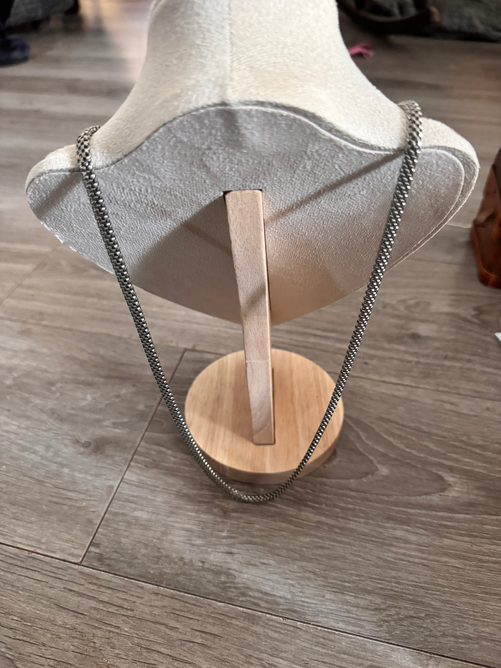 A silver chain necklace draped over a light beige fabric mannequin bust, with a small round wooden stand on a gray wood-look floor. Displaying the long length